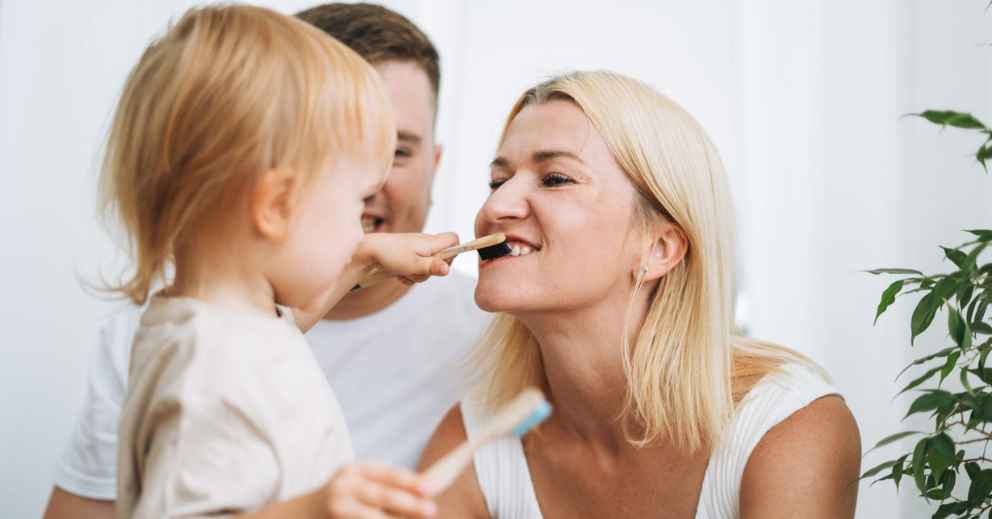 Toddler practises Oral Hygiene Tips for Children by brushing a woman's teeth as a man watches.