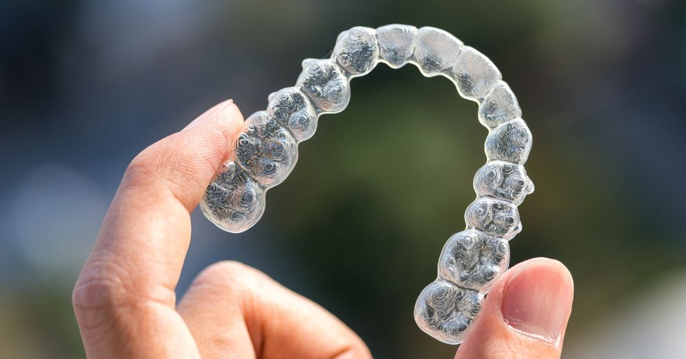 A hand holds a clear plastic dental aligner, highlighting Invisalign vs Braces options.